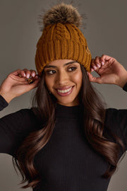 A smiling woman wears the Mustard Cable Knit Design Pom Hat by Panache Apparel Collection, paired with a black long-sleeve top, as she poses with her hands touching the hat against a plain background.