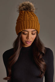 A woman with long, wavy dark hair wears the Panache Apparel Collection Mustard Cable Knit Design Pom Hat and a black ribbed turtleneck, looking downward against a plain background.