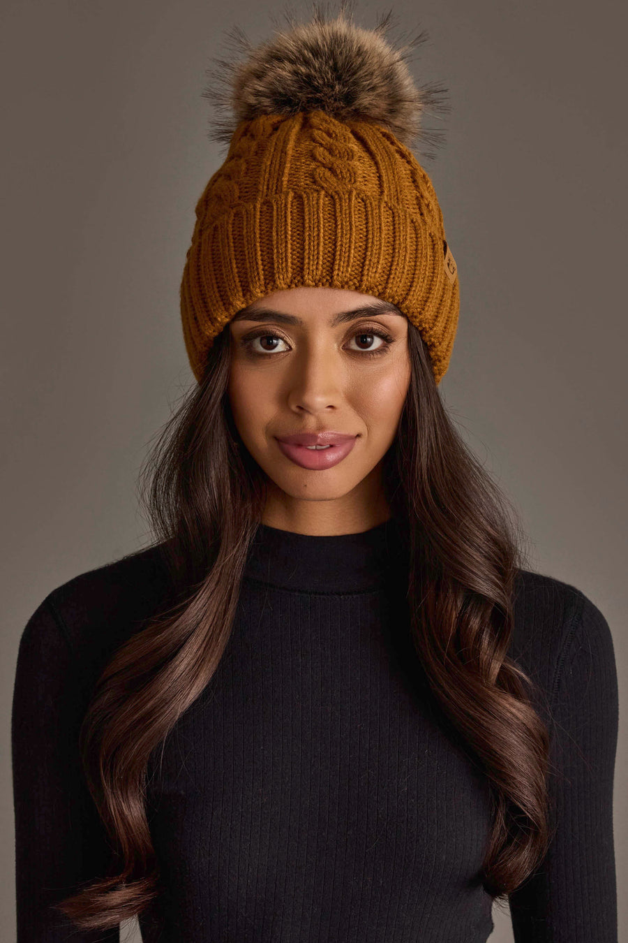 A woman with long brown hair models the Panache Apparel Collection Mustard Cable Knit Design Pom Hat, featuring a fur pom-pom, paired with a black ribbed turtleneck against a plain gray background.
