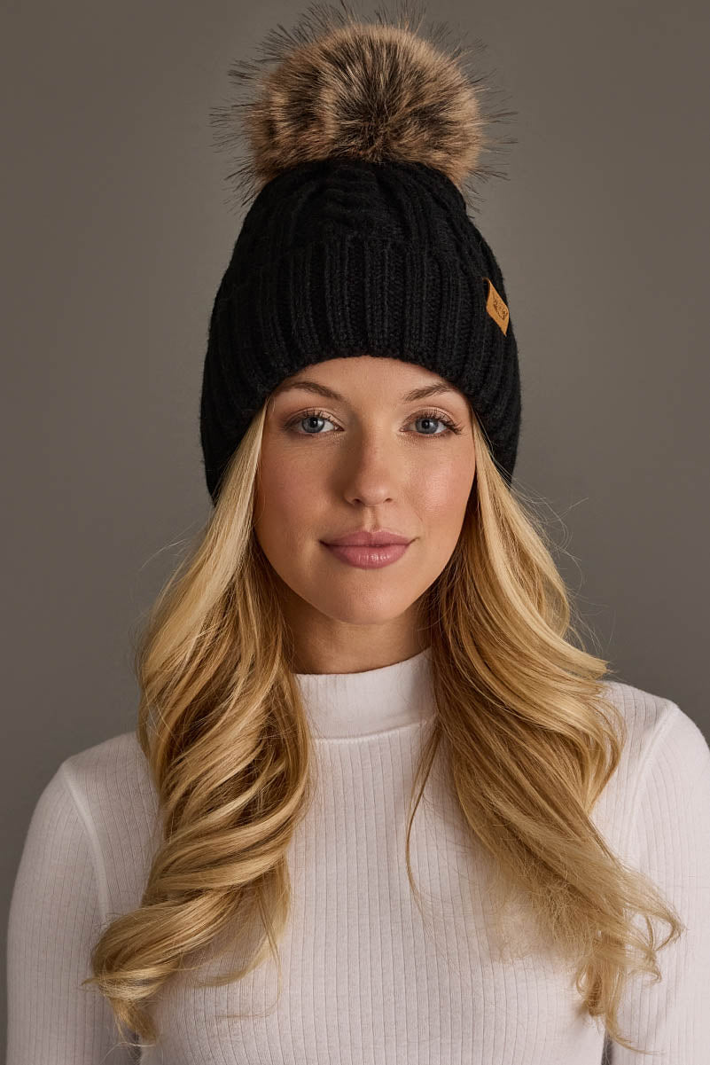 A woman with long blonde hair wears a white turtleneck sweater and the Panache Apparel Collection Black Cable Knit Design Pom Hat, featuring a large faux fur pom-pom, as she stands against a plain grey background.