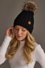 A woman with long blonde hair wears the Panache Apparel Collection Black Cable Knit Design Pom Hat, featuring a brown faux fur pom-pom, paired with a white ribbed shirt. She looks down, gently touching her hair against a neutral gray background.