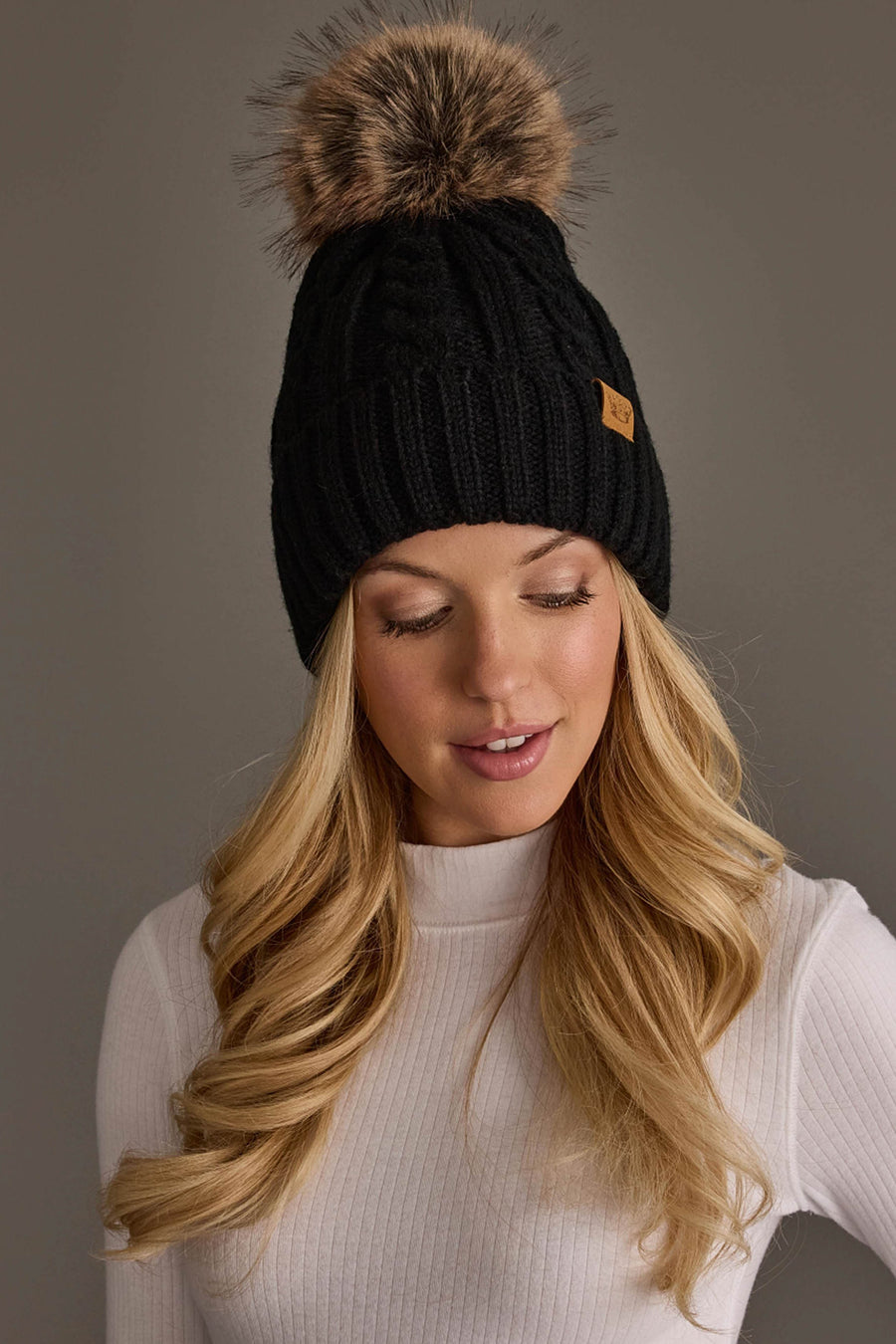 A woman with long blonde hair wears the Panache Apparel Collection Black Cable Knit Design Pom Hat and a white ribbed top, looking down against a plain gray background.