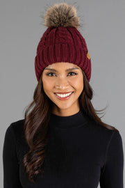 A woman wearing the Panache Apparel Collection Burgundy Cable Knit Design Pom Hat and a black long-sleeve shirt smiles against a plain gray background.