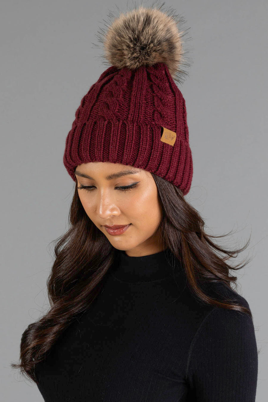A woman wearing the Panache Apparel Collection’s Burgundy Cable Knit Design Pom Hat, featuring a large faux fur pom-pom on top, looks down with a subtle smile while dressed in a black turtleneck against a plain gray background.