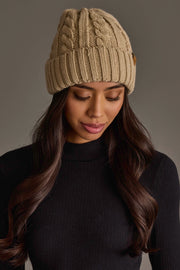 A woman with long dark hair wearing the Panache Apparel Collection’s Tan Cable Knit Design Beanie and a black ribbed turtleneck sweater, looking down with a soft expression against a plain gray background.