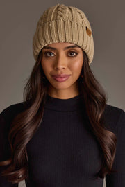 A woman with long brown hair wears the Panache Apparel Collection’s Tan Cable Knit Design Beanie and a black ribbed turtleneck sweater, looking directly at the camera against a plain gray background.