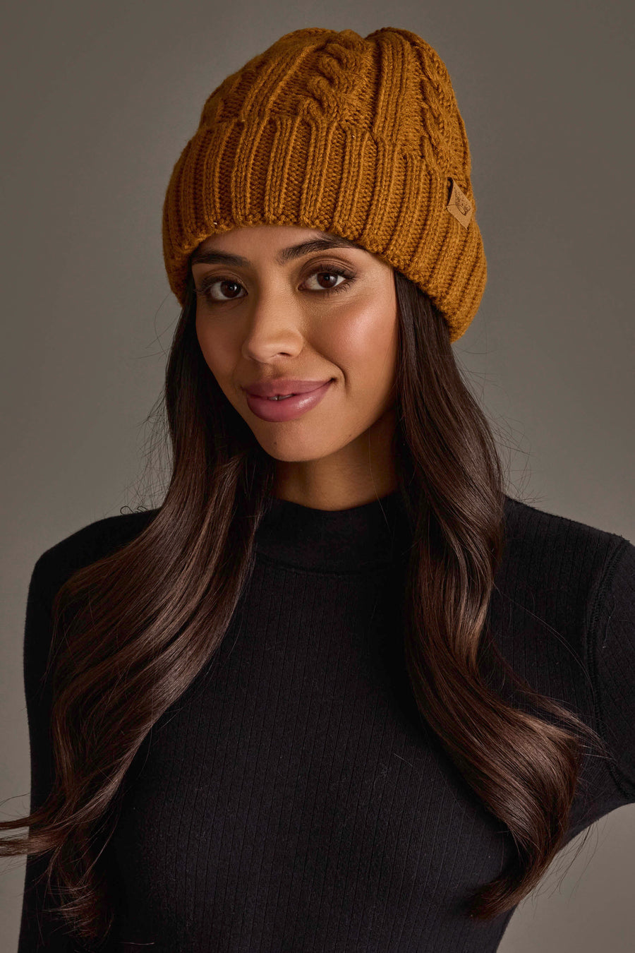 A woman with long brown hair is wearing the Panache Apparel Collection Mustard Cable Knit Design Beanie and a black ribbed sweater, smiling softly against a neutral background.