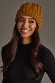 A woman with long dark hair wears the Panache Apparel Collection Mustard Cable Knit Design Beanie and a black ribbed sweater, smiling and looking slightly to the side against a plain background.