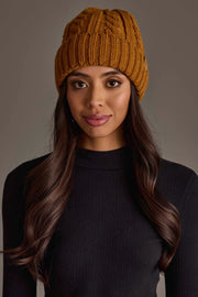 A woman with long brown hair is wearing the Panache Apparel Collection Mustard Cable Knit Design Beanie and a black ribbed sweater, standing against a plain, neutral background.
