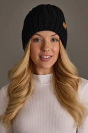 A woman with long blonde hair wears the Panache Apparel Collection Black Cable Knit Design Beanie and a white ribbed turtleneck top, smiling softly against a plain gray background.