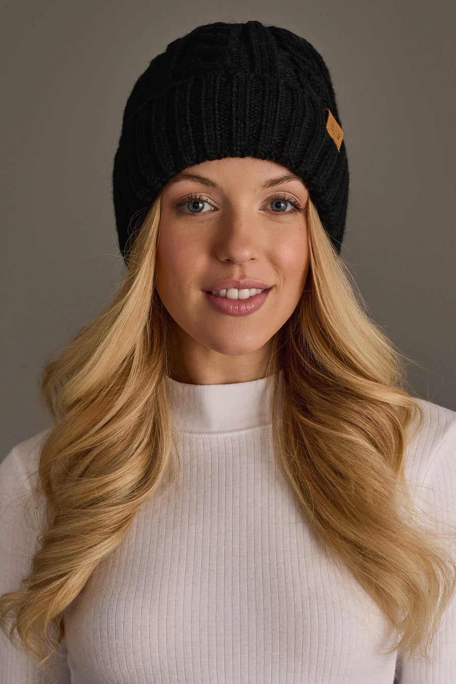 A woman with long blonde hair wears the Panache Apparel Collection Black Cable Knit Design Beanie and a white ribbed turtleneck top, smiling softly against a plain gray background.