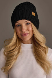 A woman with long blonde hair wears the Panache Apparel Collection Black Cable Knit Design Beanie and a white ribbed turtleneck sweater, smiling softly against a neutral background.