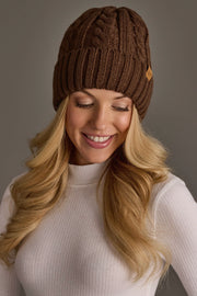A woman with long blonde hair is smiling and looking down against a plain gray background, wearing the Panache Apparel Collection Brown Cable Knit Design Beanie and a white ribbed turtleneck sweater.