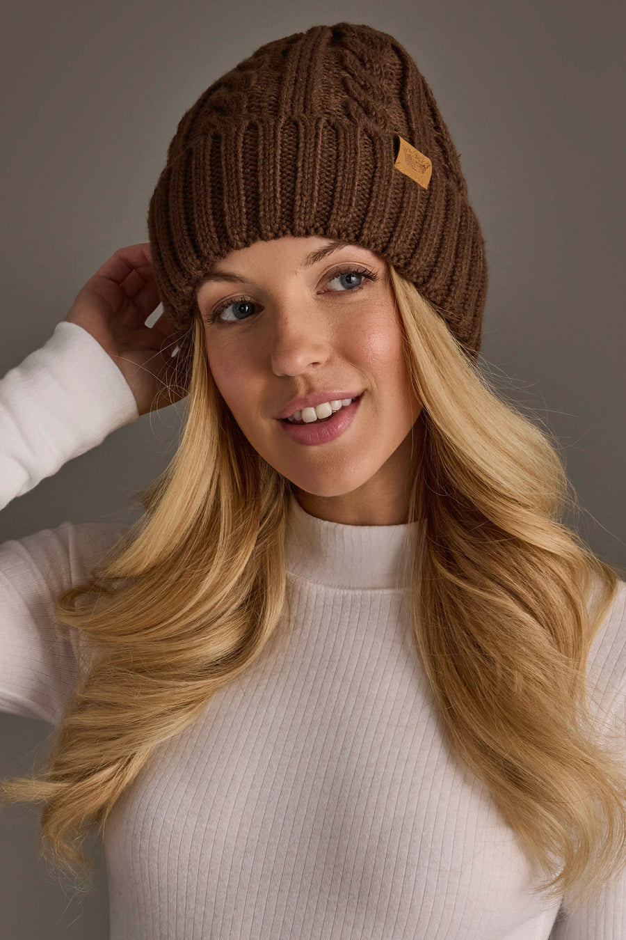 A woman with long blonde hair smiles and touches the side of her Brown Cable Knit Design Beanie from Panache Apparel Collection, wearing a white ribbed shirt against a neutral background.