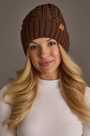 Wearing the Panache Apparel Collection’s Brown Cable Knit Design Beanie and a white ribbed sweater, a person with long blonde hair smiles softly against a neutral background.