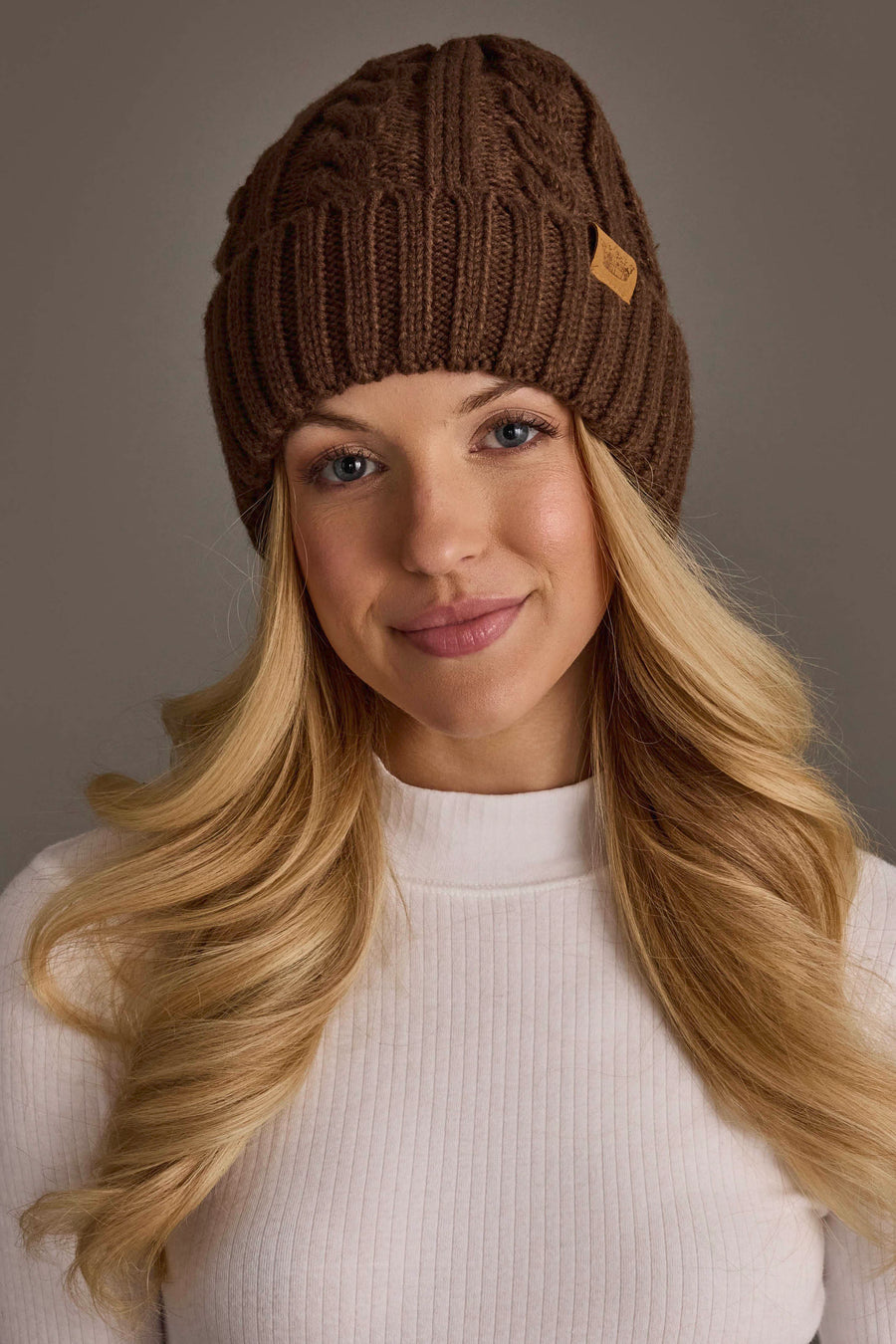 Wearing the Panache Apparel Collection’s Brown Cable Knit Design Beanie and a white ribbed sweater, a person with long blonde hair smiles softly against a neutral background.