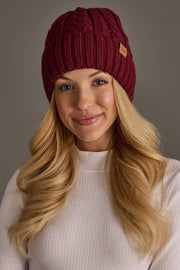 A smiling woman with long blonde hair models the Panache Apparel Collection Burgundy Cable Knit Design Beanie and a white ribbed sweater, standing against a plain gray background.