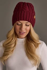 A woman with long blonde hair wears the Panache Apparel Collection Burgundy Cable Knit Design Beanie and a white ribbed sweater, smiling softly as she looks down against a neutral background.