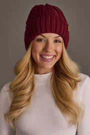 A smiling woman with long blonde hair wears the Burgundy Cable Knit Design Beanie from Panache Apparel Collection and a white ribbed turtleneck sweater, standing against a plain gray background.