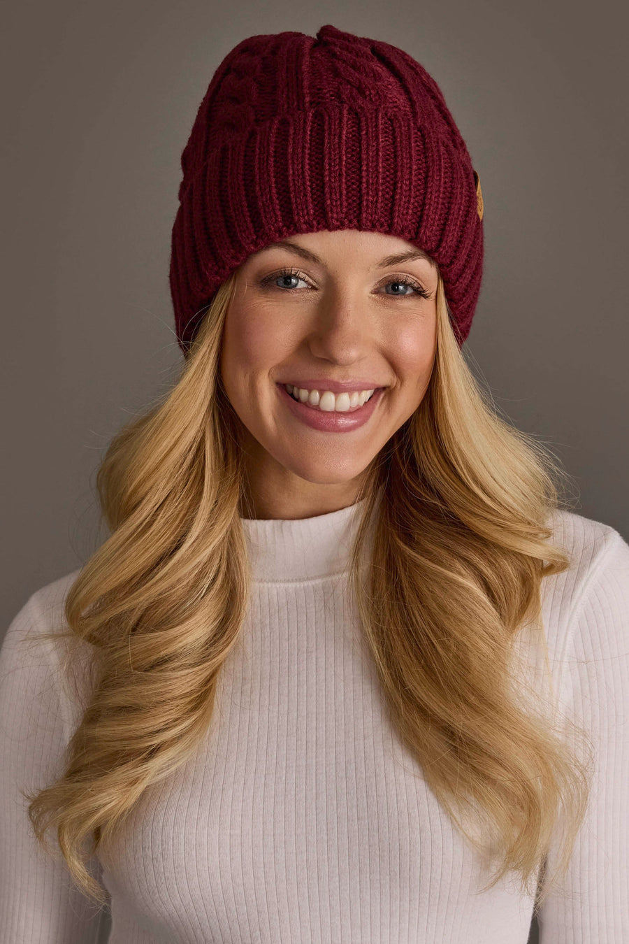 A smiling woman with long blonde hair wears the Burgundy Cable Knit Design Beanie from Panache Apparel Collection and a white ribbed turtleneck sweater, standing against a plain gray background.