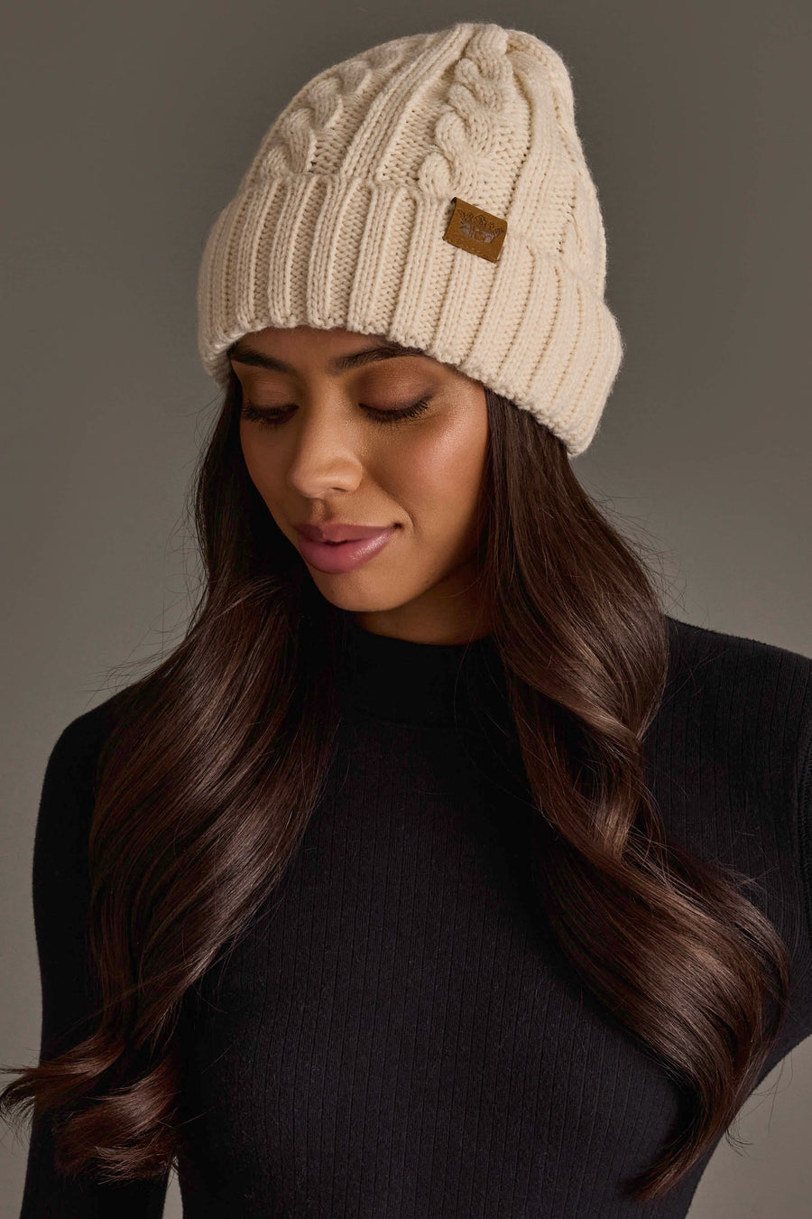 A woman with long, wavy brown hair wears the Cream Cable Knit Design Beanie from Panache Apparel Collection and a black ribbed sweater, looking down with a neutral expression against a plain gray background.