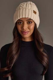 A woman with long brown hair wears the Cream Cable Knit Design Beanie from Panache Apparel Collection and a black ribbed sweater, standing against a neutral background and looking at the camera with a slight smile.
