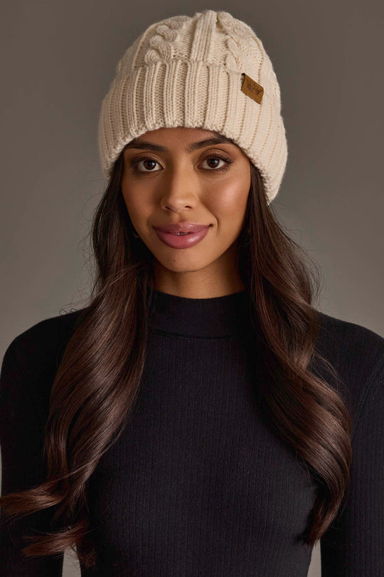 A woman with long brown hair wears the Cream Cable Knit Design Beanie from Panache Apparel Collection and a black ribbed sweater, standing against a neutral background and looking at the camera with a slight smile.