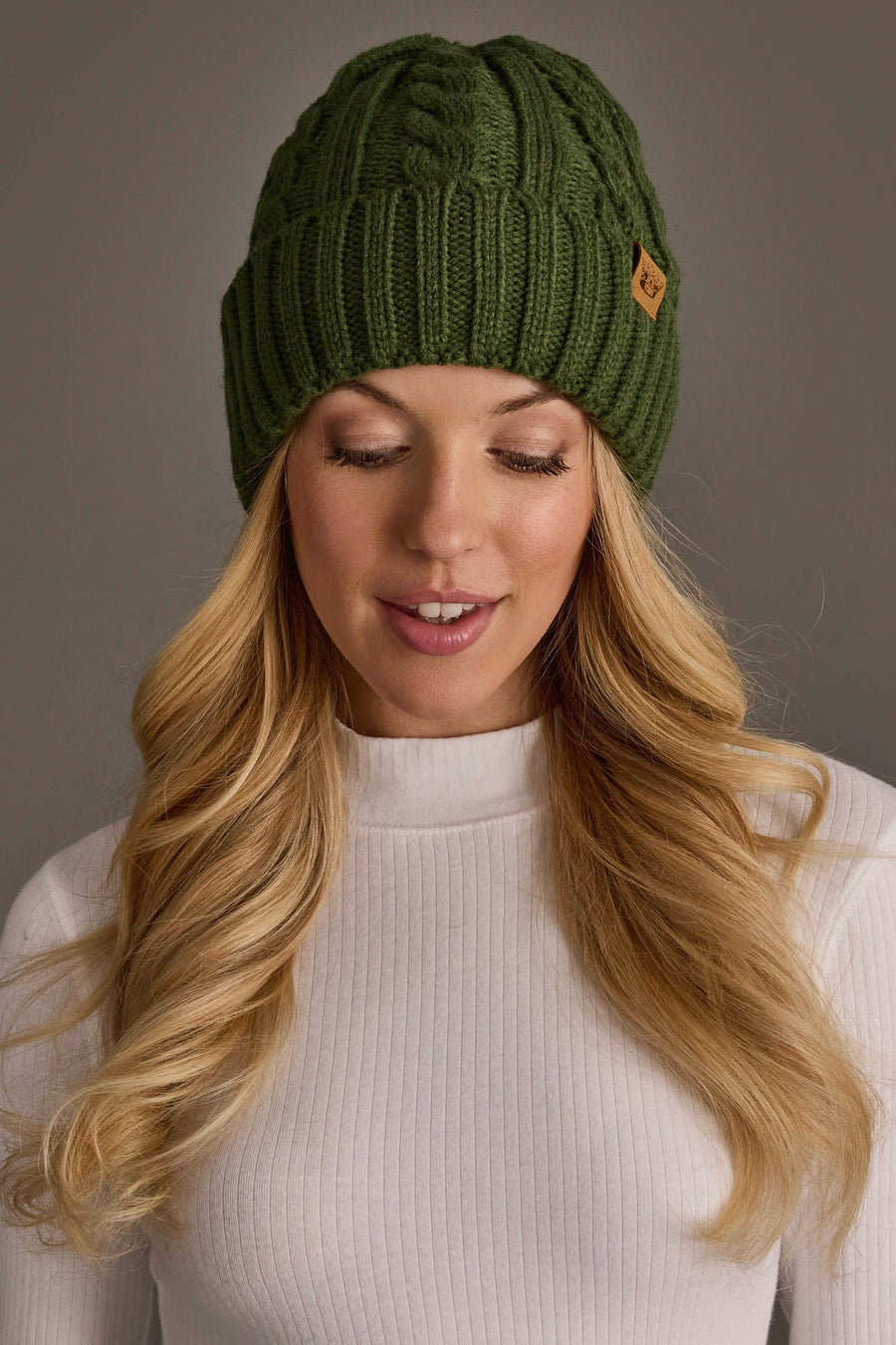 A woman with long blonde hair wears the Panache Apparel Collection’s Dark Green Cable Knit Design Beanie and a white ribbed turtleneck, looking down and smiling softly against a neutral gray background.