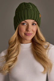 A woman with long blonde hair wears a Panache Apparel Collection Dark Green Cable Knit Design Beanie and a white ribbed turtleneck, smiling softly against a neutral background.
