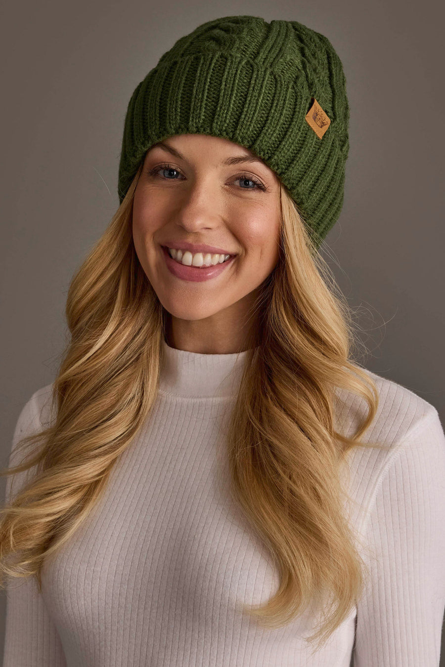 A woman with long blonde hair wears the Panache Apparel Collection Dark Green Cable Knit Design Beanie and a white ribbed sweater, smiling at the camera against a neutral background.