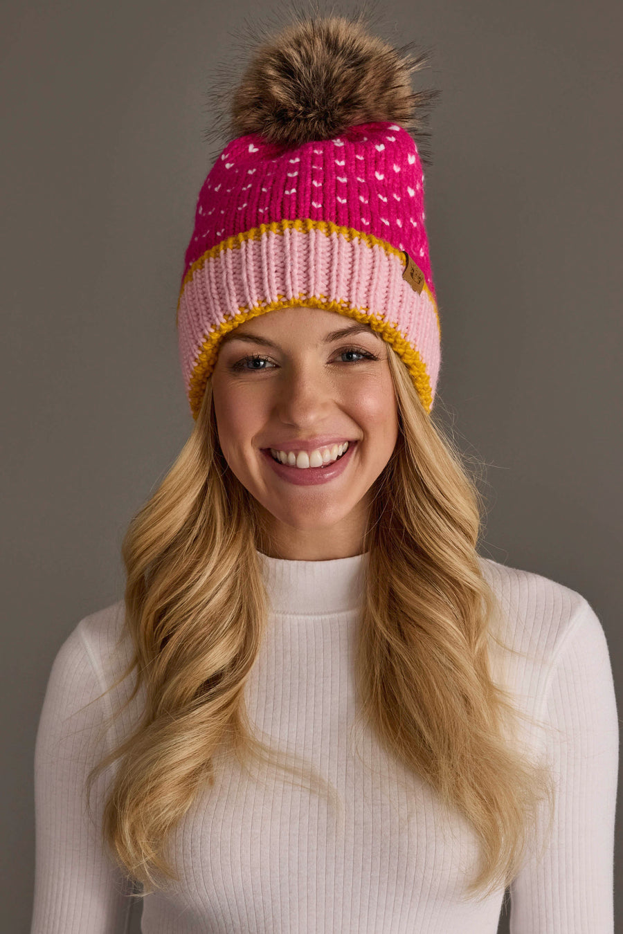 A smiling woman with long blonde hair wears the Panache Apparel Collection Pink, White & Light Pink Heart Design Pom Hat and a white ribbed sweater, set against a plain gray background.