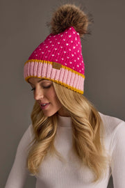A woman with long blonde hair wears the Panache Apparel Collection’s Pink, White & Light Pink Heart Design Pom Hat, paired with a white ribbed turtleneck sweater. She is looking down and smiling softly.