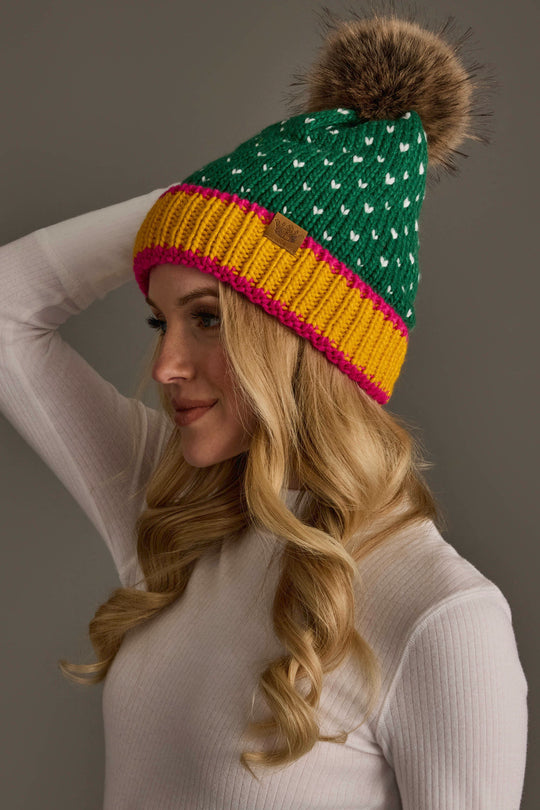 A woman wearing the Panache Apparel Collection’s Green, White & Mustard Heart Design Pom Hat, a green knit beanie featuring white heart designs, a yellow and pink rim, and a large brown faux fur pom-pom, pairs it with a white ribbed turtleneck sweater. She looks to the side against a plain background.