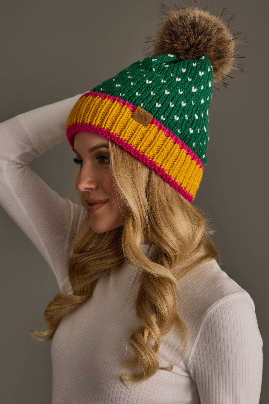 A woman wearing the Panache Apparel Collection’s Green, White & Mustard Heart Design Pom Hat, a green knit beanie featuring white heart designs, a yellow and pink rim, and a large brown faux fur pom-pom, pairs it with a white ribbed turtleneck sweater. She looks to the side against a plain background.