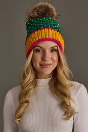 A smiling woman with long blonde hair wears the Green, White & Mustard Heart Design Pom Hat by Panache Apparel Collection, featuring a colorful knit pattern and a fur pom-pom, paired with a white ribbed turtleneck.