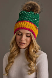 A woman with long blonde hair wears the Panache Apparel Collection’s Green, White & Mustard Heart Design Pom Hat, featuring white heart details, a large brown pom-pom, and a yellow and pink band. She pairs it with a white ribbed sweater and looks down with a slight smile against a plain background.