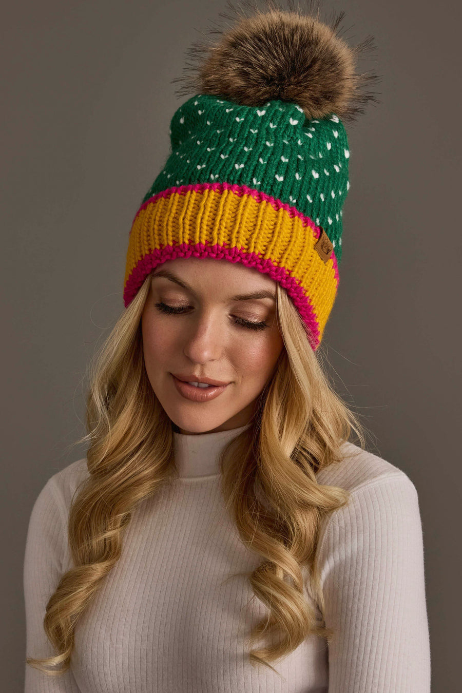A woman with long blonde hair wears the Panache Apparel Collection’s Green, White & Mustard Heart Design Pom Hat, featuring white heart details, a large brown pom-pom, and a yellow and pink band. She pairs it with a white ribbed sweater and looks down with a slight smile against a plain background.