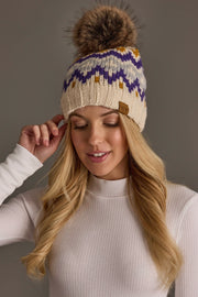 A woman with long blonde hair smiles softly with her eyes closed, wearing a Cream, Gold, Purple & Gray Patterned Pom Hat from Panache Apparel Collection and a white ribbed top.