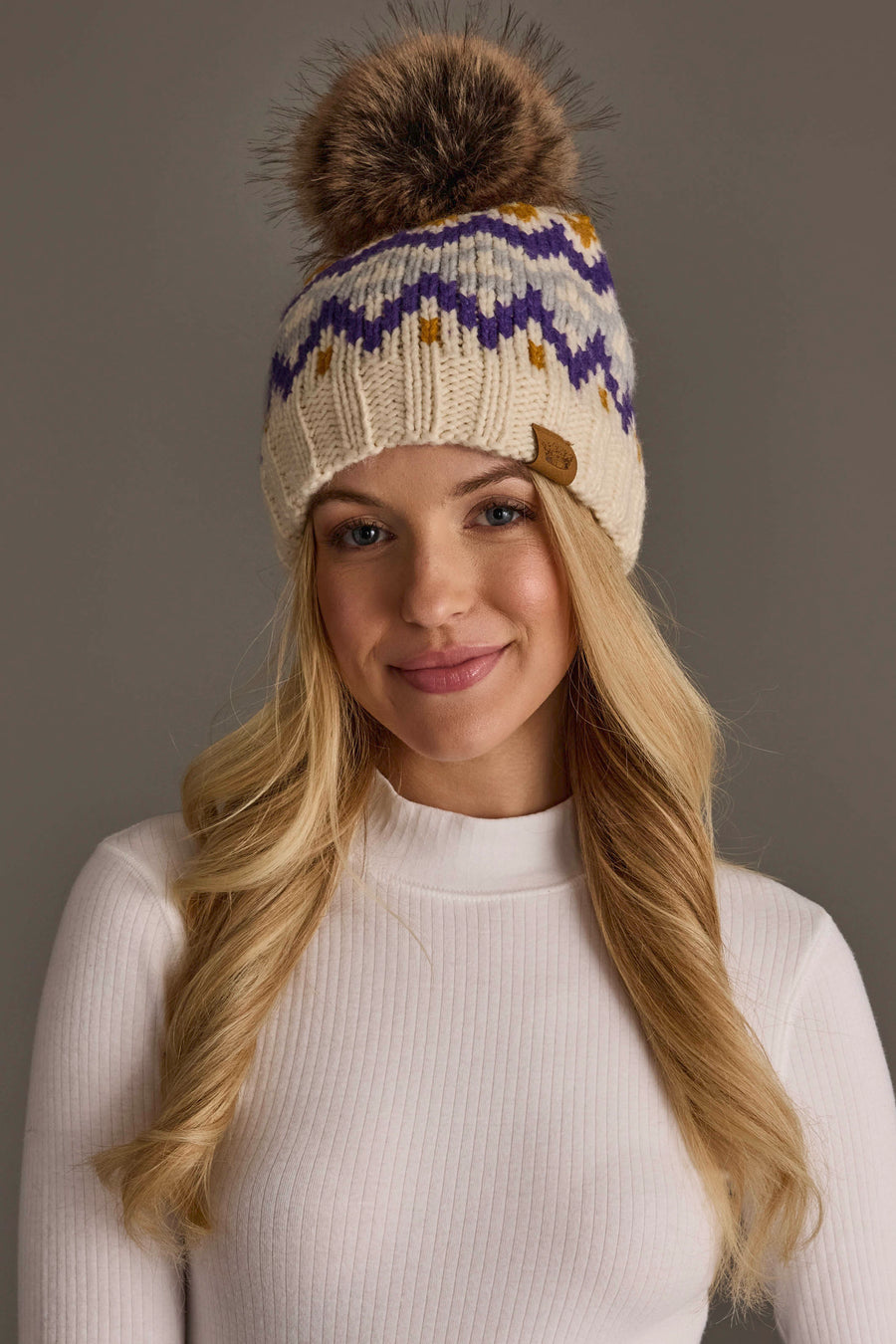A woman smiles against a plain gray background, wearing a white long-sleeve shirt and the Cream, Gold, Purple & Gray Patterned Pom Hat from Panache Apparel Collection.