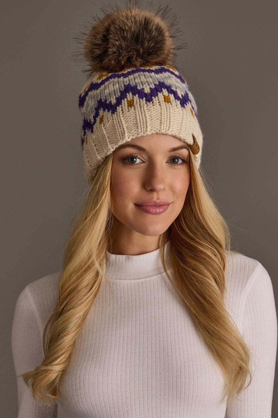 A woman with long blonde hair models the Panache Apparel Collection’s Cream, Gold, Purple & Gray Patterned Pom Hat featuring a knit design with a fur pom-pom paired with a white ribbed sweater against a neutral background.
