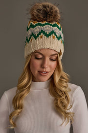 A woman with long blonde hair wears the Panache Apparel Collection Cream, Gold, Green & Gray Patterned Pom Hat, featuring a large fur pom-pom. She is looking down and dressed in a white ribbed turtleneck top against a plain gray background.