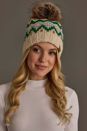 A woman with long blonde hair wears the Cream, Gold, Green & Gray Patterned Pom Hat by Panache Apparel Collection a knit beanie featuring a fur pom-pom and an eye-catching pattern. She is dressed in a white ribbed top and smiles softly against a plain background.