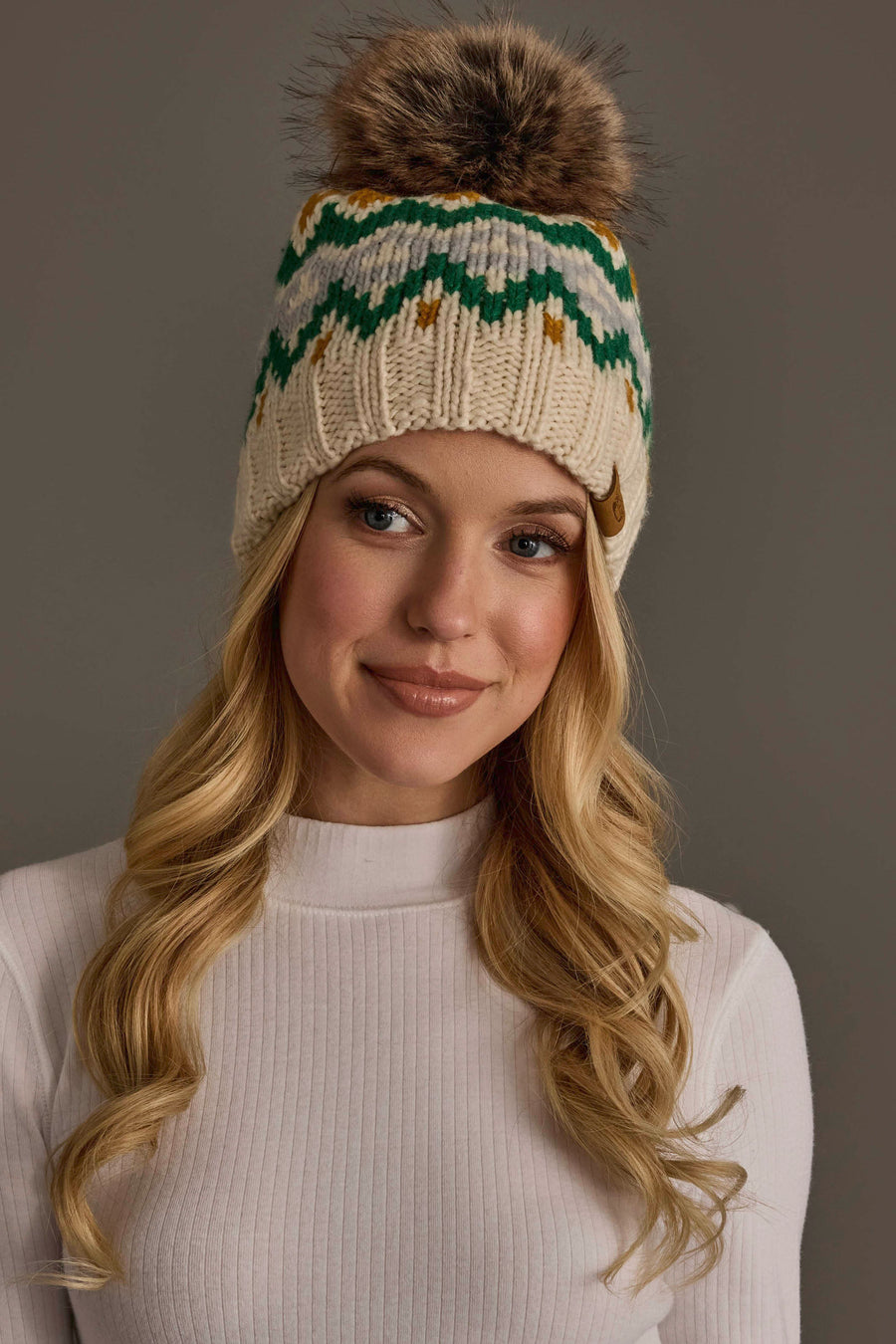 A woman with long blonde hair wears the Cream, Gold, Green & Gray Patterned Pom Hat by Panache Apparel Collection a knit beanie featuring a fur pom-pom and an eye-catching pattern. She is dressed in a white ribbed top and smiles softly against a plain background.