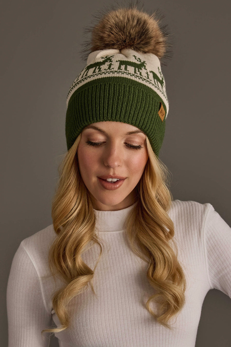 An Olive & Beige Deer Patterned Pom Hat with a green ribbed brim, fleece lining, cream-colored top featuring a green deer pattern, and a large faux fur pom-pom.