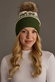A person wears a fleece-lined Olive & Beige Deer Patterned Pom Hat by Panache Apparel Collection, featuring a deer pattern and a large brown faux-fur pom-pom.