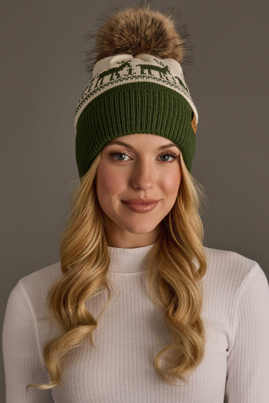 A person wears a fleece-lined Olive & Beige Deer Patterned Pom Hat by Panache Apparel Collection, featuring a deer pattern and a large brown faux-fur pom-pom.