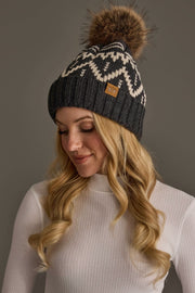 A woman with long blonde hair wears a white turtleneck and the Charcoal & Beige Patterned Pom Hat from Panache Apparel Collection, looking down and smiling.