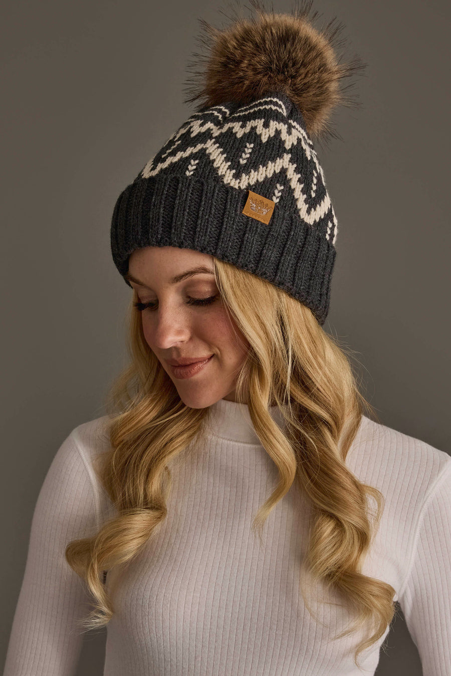 A woman with long blonde hair wears a white turtleneck and the Charcoal & Beige Patterned Pom Hat from Panache Apparel Collection, looking down and smiling.