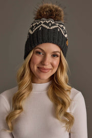 A woman with long blonde hair, smiling at the camera against a plain background, wears a white ribbed top and the Panache Apparel Collection Charcoal & Beige Patterned Pom Hat.
