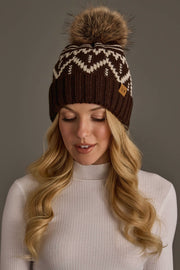 A woman with long blonde hair wears the Chocolate Brown & Beige Patterned Pom Hat from Panache Apparel Collection, featuring a white design. She is looking down and wearing a white ribbed turtleneck sweater.